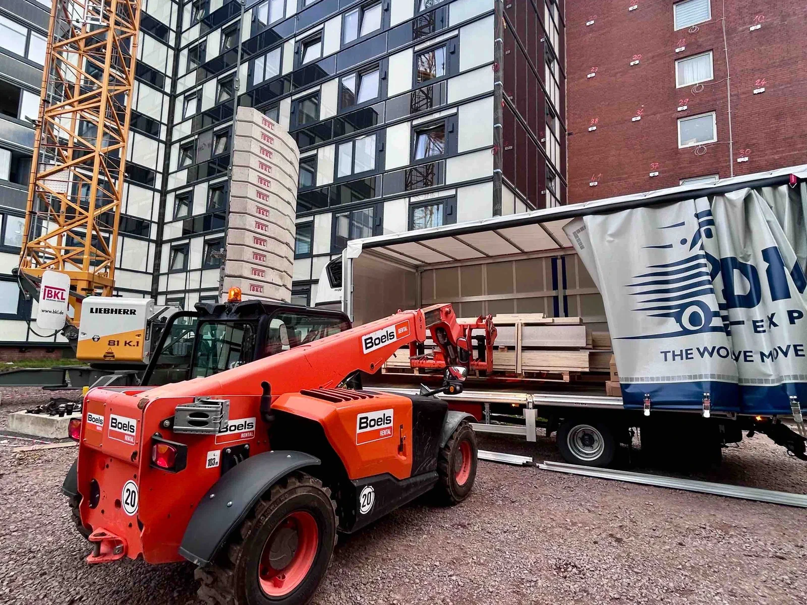 A telehandler loading a pallet through the open side of a DMF Express curtain-side truck on a construction site